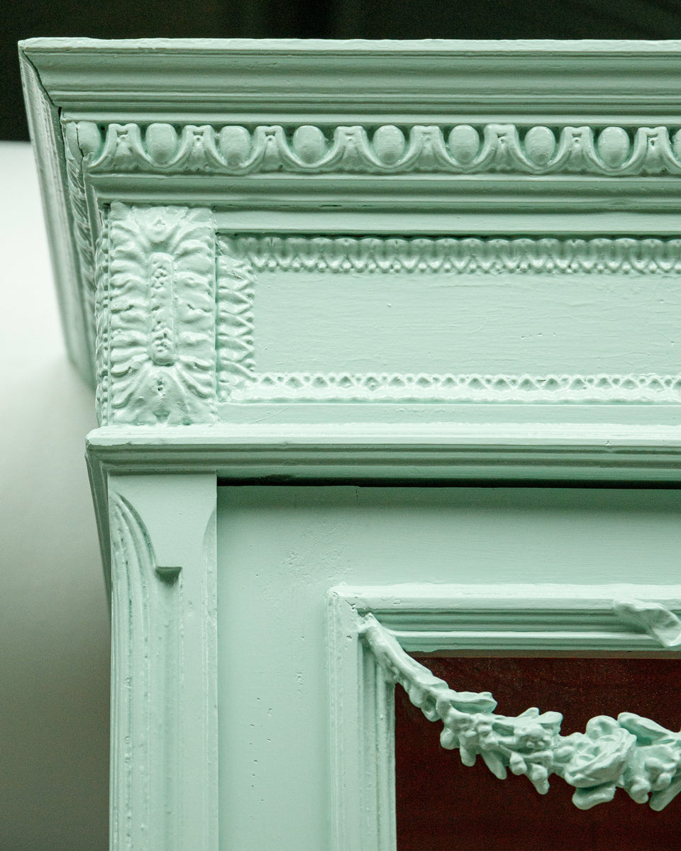 Close-up of the left upper corner of a French Louis XVI-style vitrine cabinet with a soft mint green finish, glass doors, and contrasting red interior, intricate detailing.