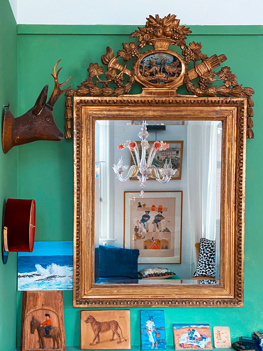 Antique French gilt mirror with an ornate carved crest featuring a La Fontaine fable scene, mounted on a green wall. The mirror reflects a Murano glass chandelier with pink flowers. Below the mirror is a colorful arrangement of small paintings and wooden carvings, with a mounted deer head and red velvet hat on the wall.