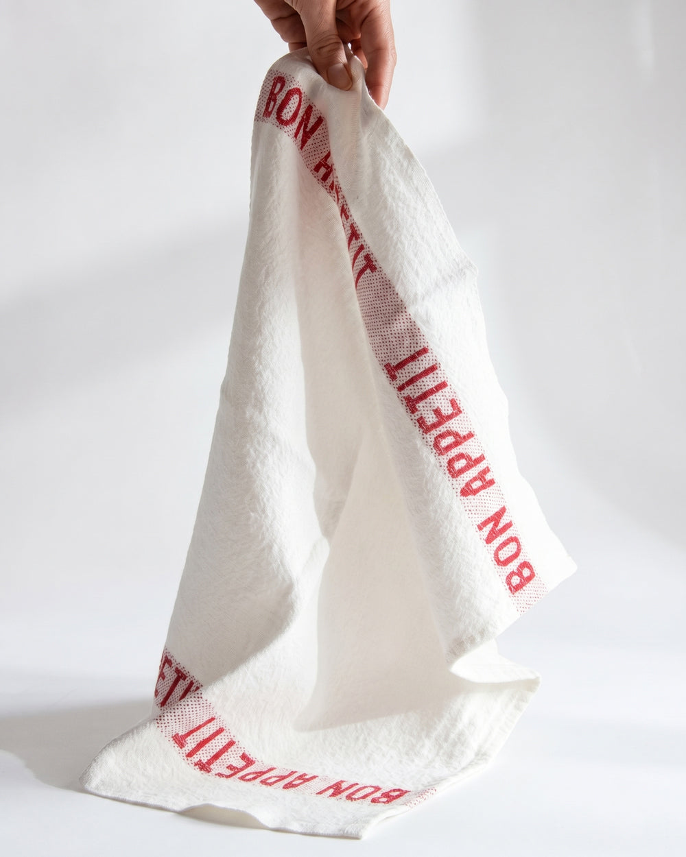 Hand holding a white linen napkin with red text Bon Appetit. By Charvet Editions, available at Wildschut Antiques.