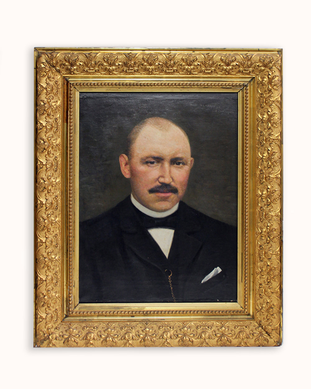 A framed antique portrait oil painting of a man with a mustache, wearing a dark suit and white shirt, enclosed in an ornate gold frame against a light background. Available via Wildschut Antiques.