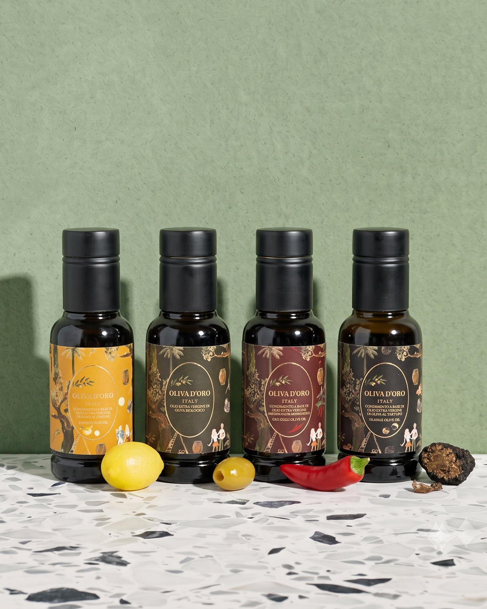 Four bottles of olive oil from the brand Oliva d'Oro, arranged on a white terrazzo surface against a textured sage green background. The bottles are styled with fresh ingredients including a lemon, a green olive, a red chili pepper, and a piece of black truffle. Available via the Pantry Collection from Wildschut Antiques.
