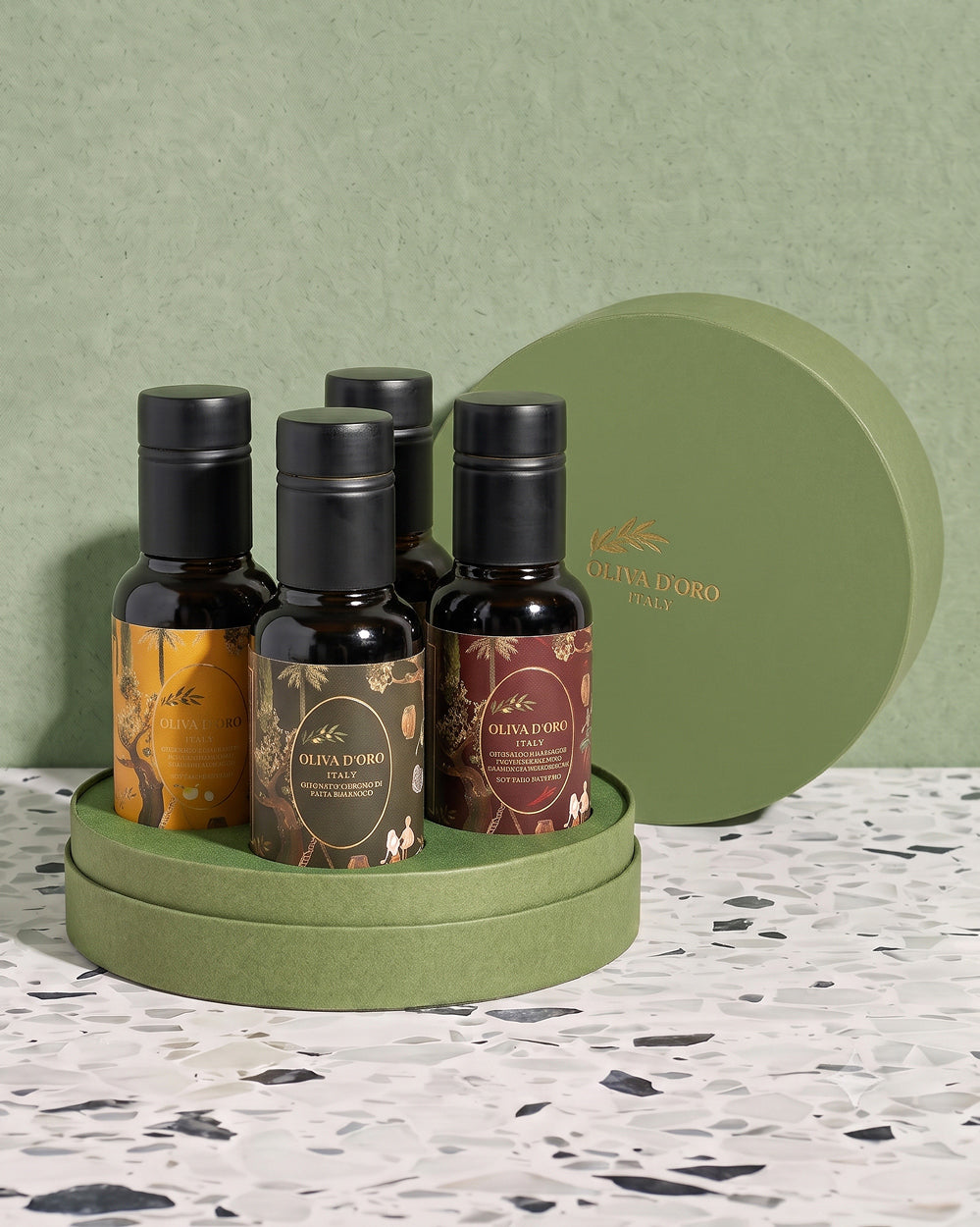 Four small olive oil bottles with black caps and decorative labels are arranged in a round green gift box on a matching round tray, with the lid leaning behind them. The setting includes a textured sage green wall and a speckled white surface.