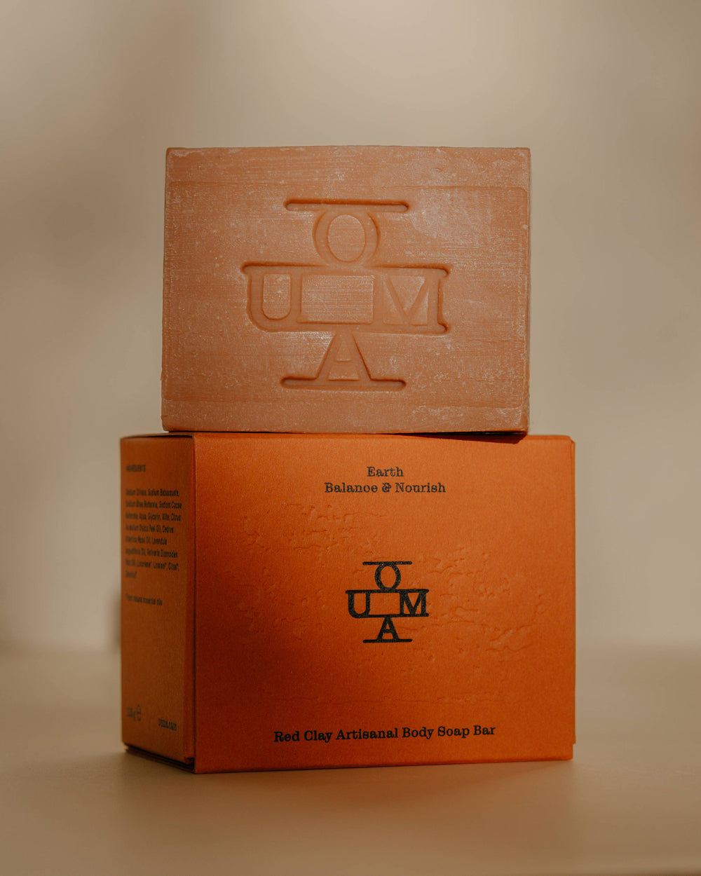A rectangular brown-red-colored soap bar with an embossed OUMA logo, positioned on top of its textured red packaging box labeled 'Earth Balance & Nourish'.