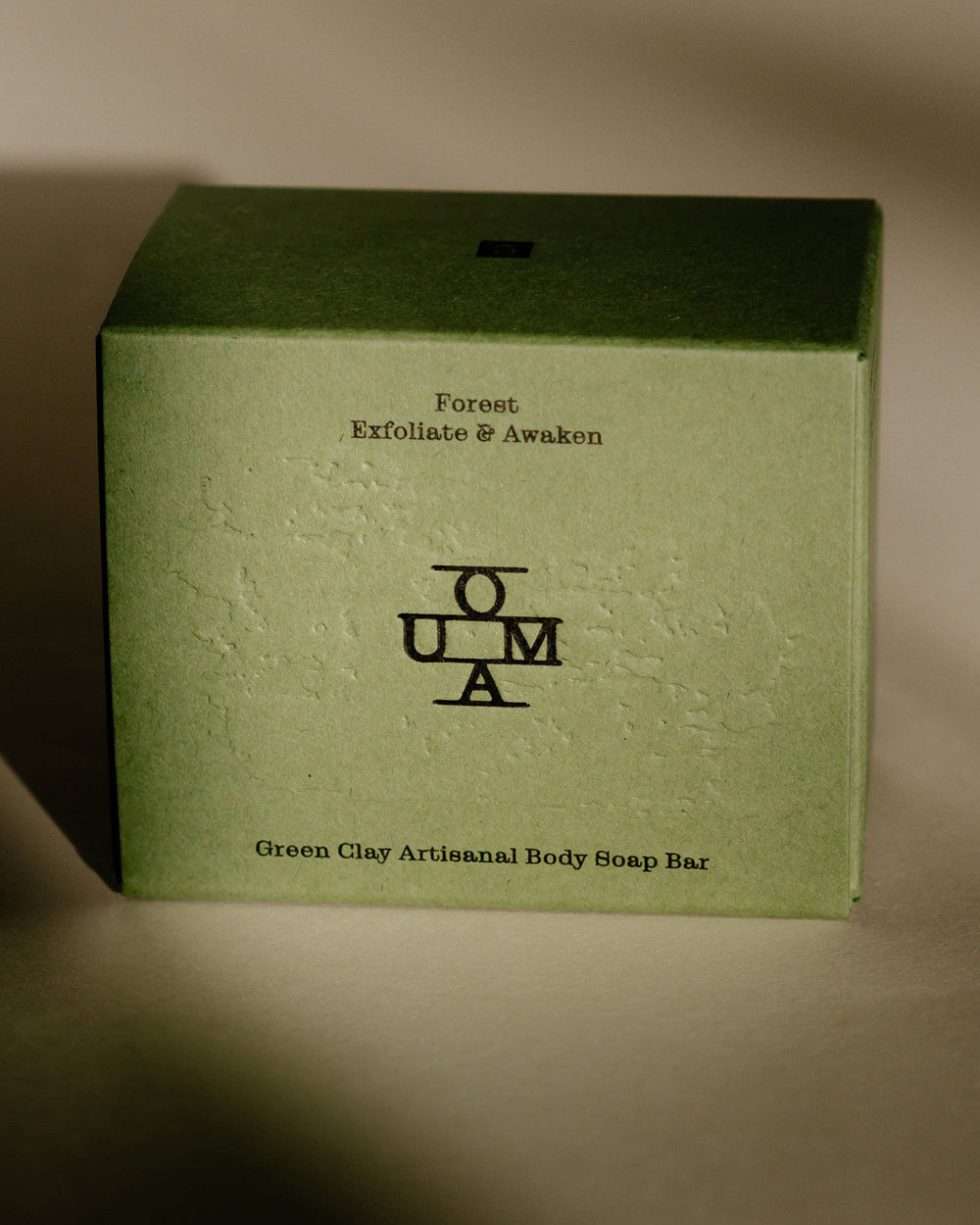 A square green cardboard box for a body soap bar, with black text reading 'Forest Exfoliate & Awaken' and the OUMA logo centered on the front, photographed against a plain beige background. Available at Wildschut Antiques & Decor.