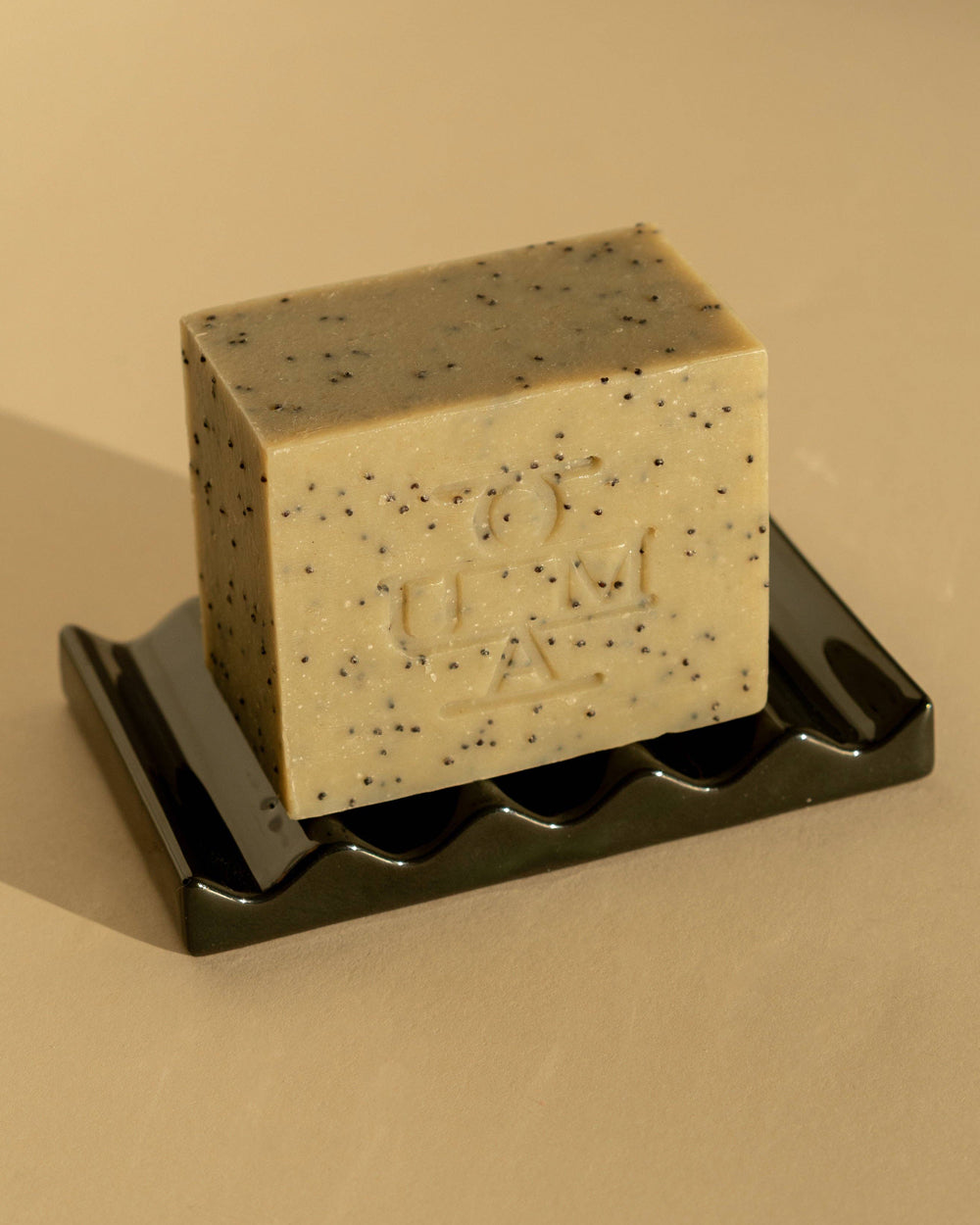 A rectangular bar of soap standing on a ceramic dish, photographed against a plain light beige background. The soap has a muted sage-green tone with scattered dark specks and an embossed geometric logo on the front that reads OUMA.