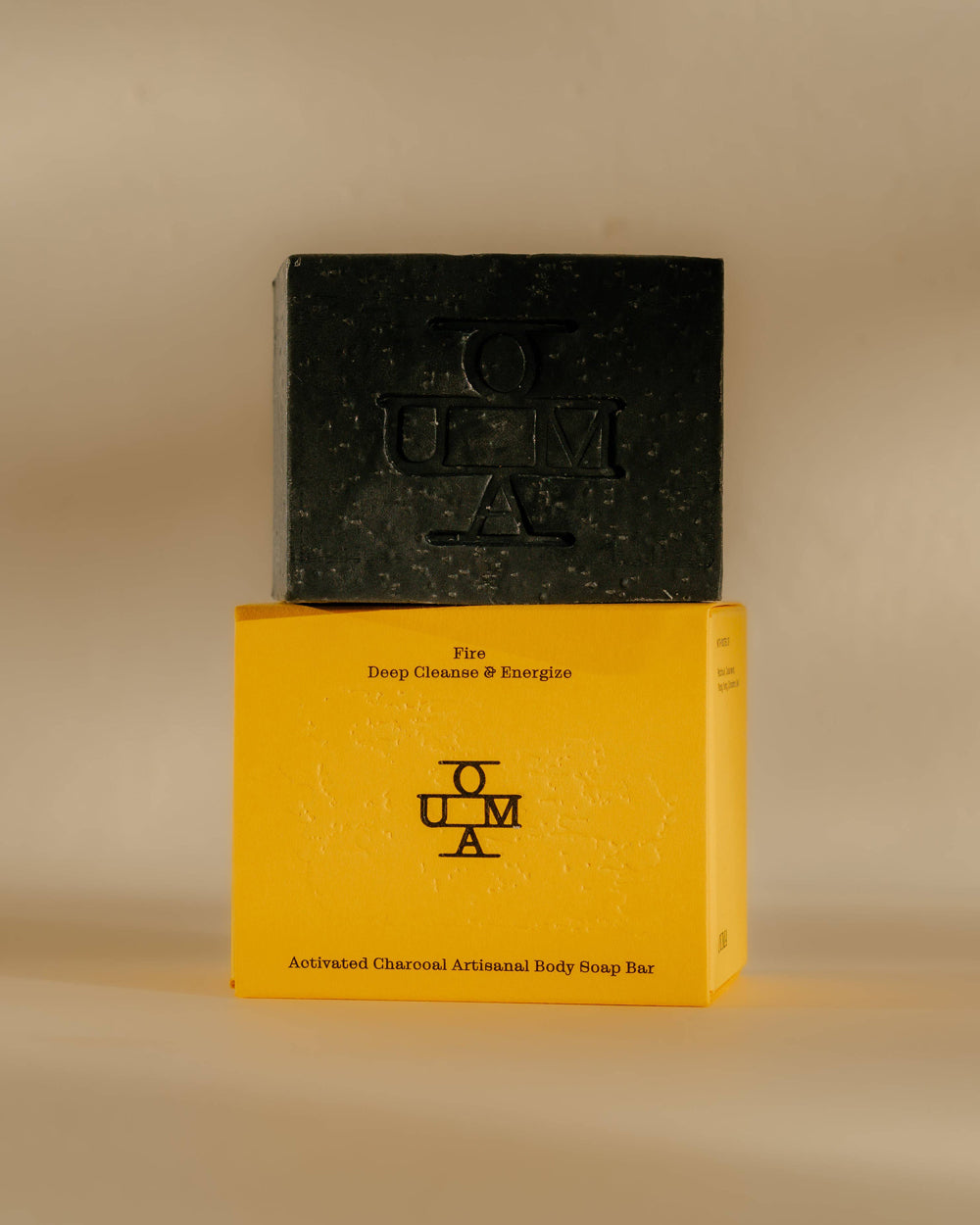 A dark charcoal body soap bar stacked on top of its bright yellow packaging box, set against a warm beige background. The brand name 'OUMA' is embossed on the soap and printed on the box. Available at Wildschut Antiques & Decor.