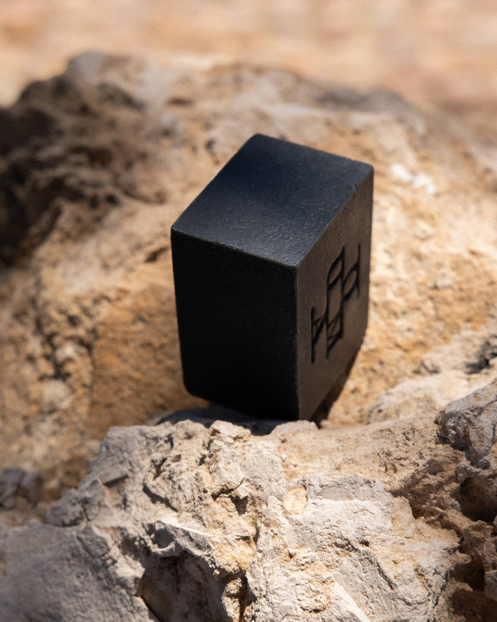 A matte black cube-shaped block of soap with an embossed logo on one side, resting on textured tan and gray rocks outdoors.