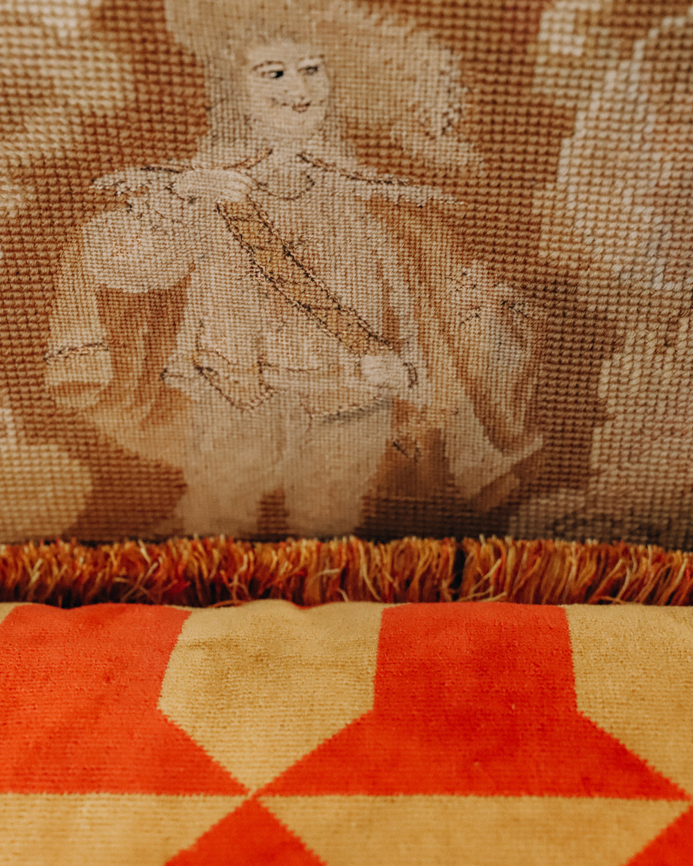 Close-up of a decorative pillow with a yellow and red pattern and fringed edges, set against a chair upholstered in Aubusson tapestry depicting a man in an elegant classical costume and a feathered hat.