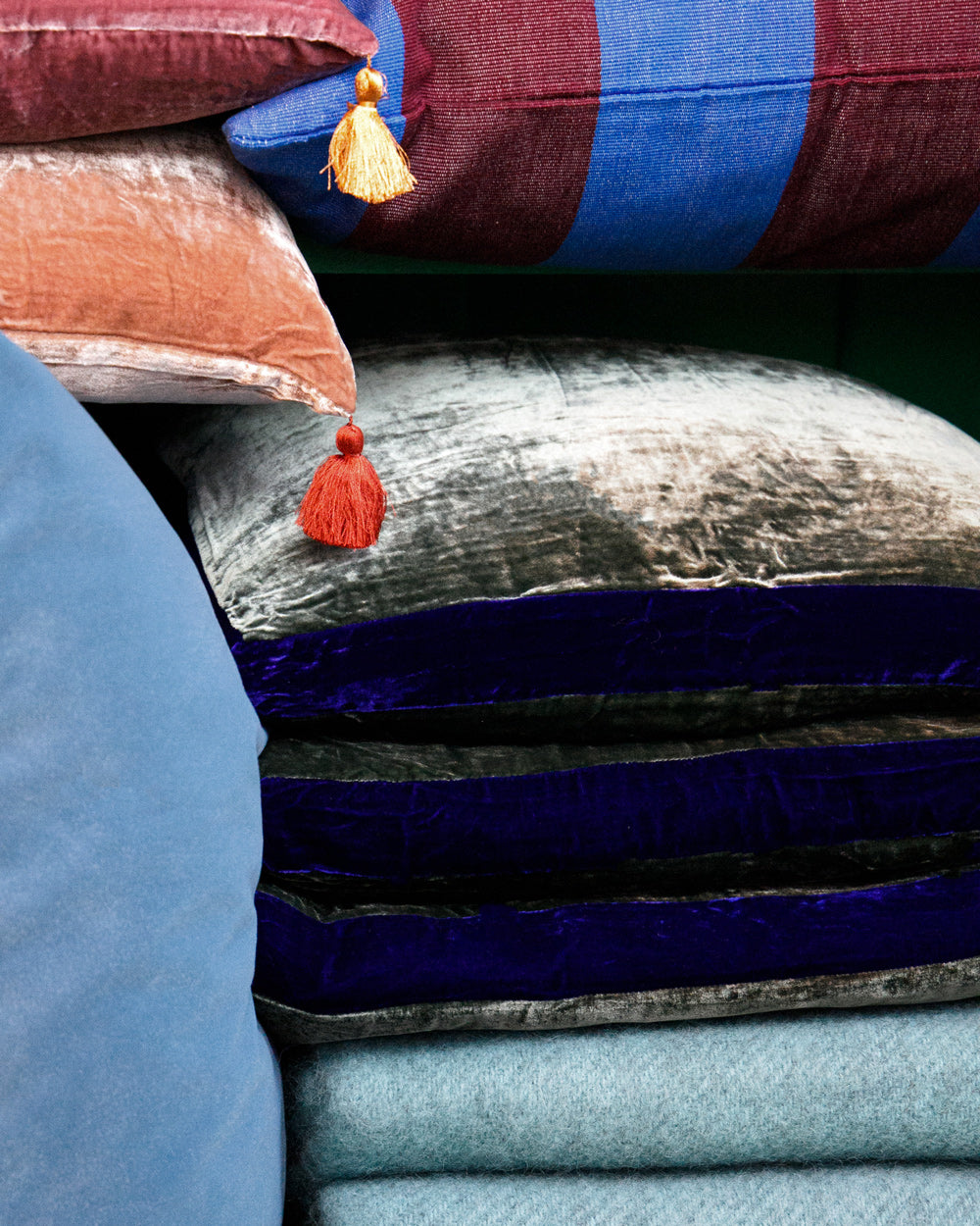 Stack of colourful cushions featuring blue and aubergine striped designs, silk velvet velour cushions with contrasting borders, and tasselled cushions, styled on turquoise blue wool blankets. Designed by Monde Sauvage and available at Wildschut Antiques.