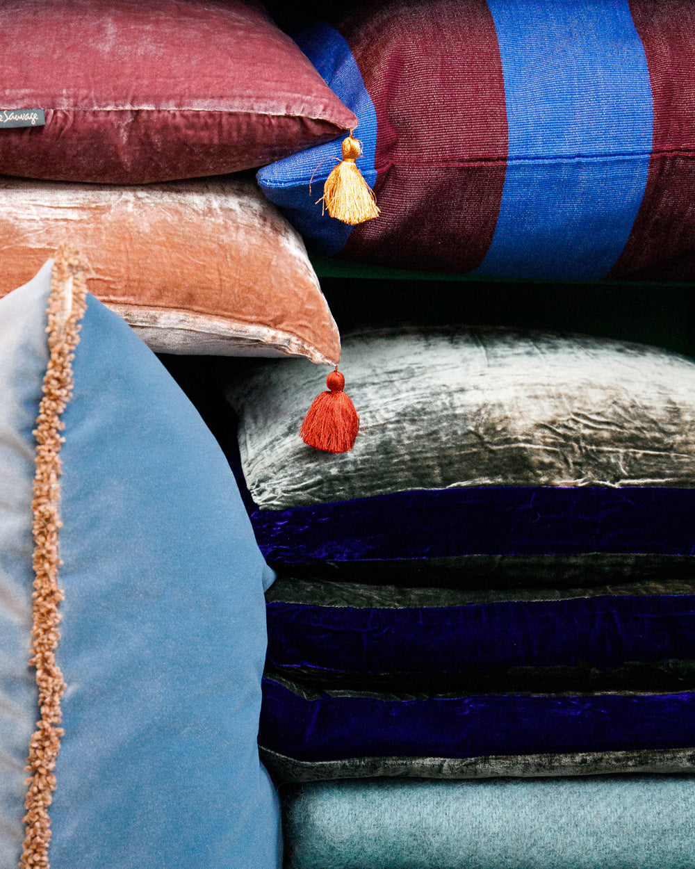 Stack of colourful cushions featuring blue and aubergine striped designs, silk velvet velour cushions with contrasting borders, and tasselled cushions, styled on turquoise blue wool blankets. Designed by Monde Sauvage and available at Wildschut Antiques.
