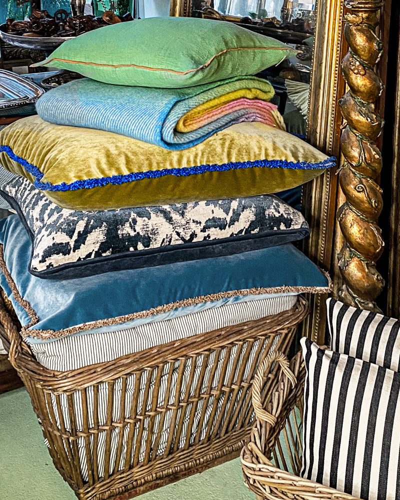 Stack of colorful pillows in a wicker basket with decorative antique mirrors in the background, available via Wildschut Antiques