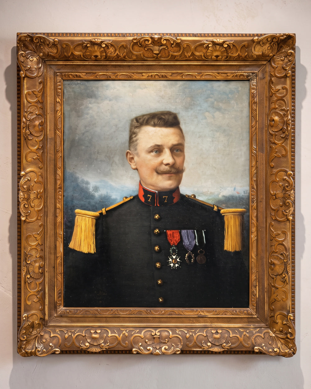 A framed historical portrait oil painting of a military officer in a dark uniform with gold epaulets and medals, set within an ornate carved gold frame against a light wall. Available at Wildschut Antiques.