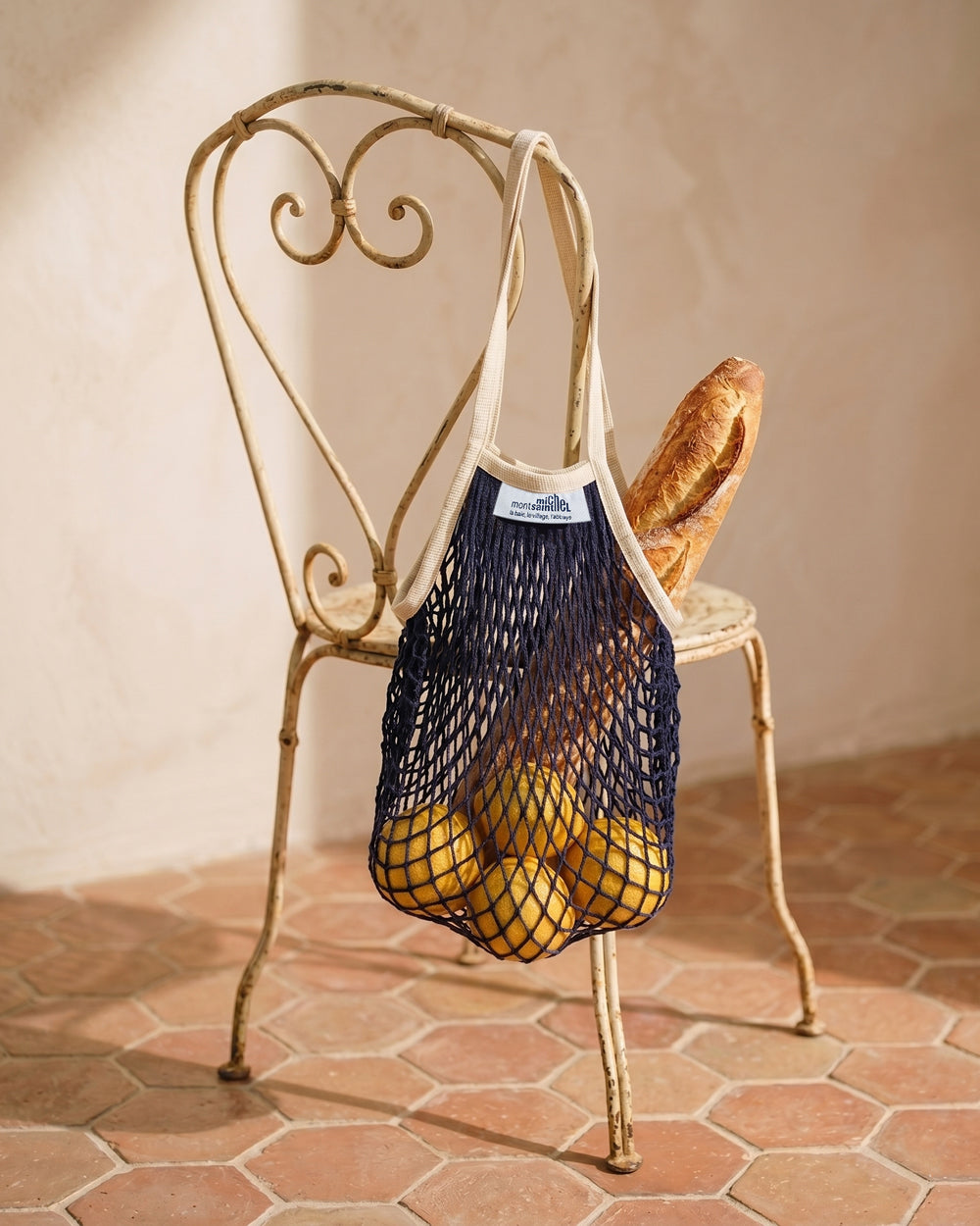 Net shopping bag with bread and grapefruits on a decorative vintage French metal chair against a beige wall. By Filt 1860, available at Wildschut Antiques.