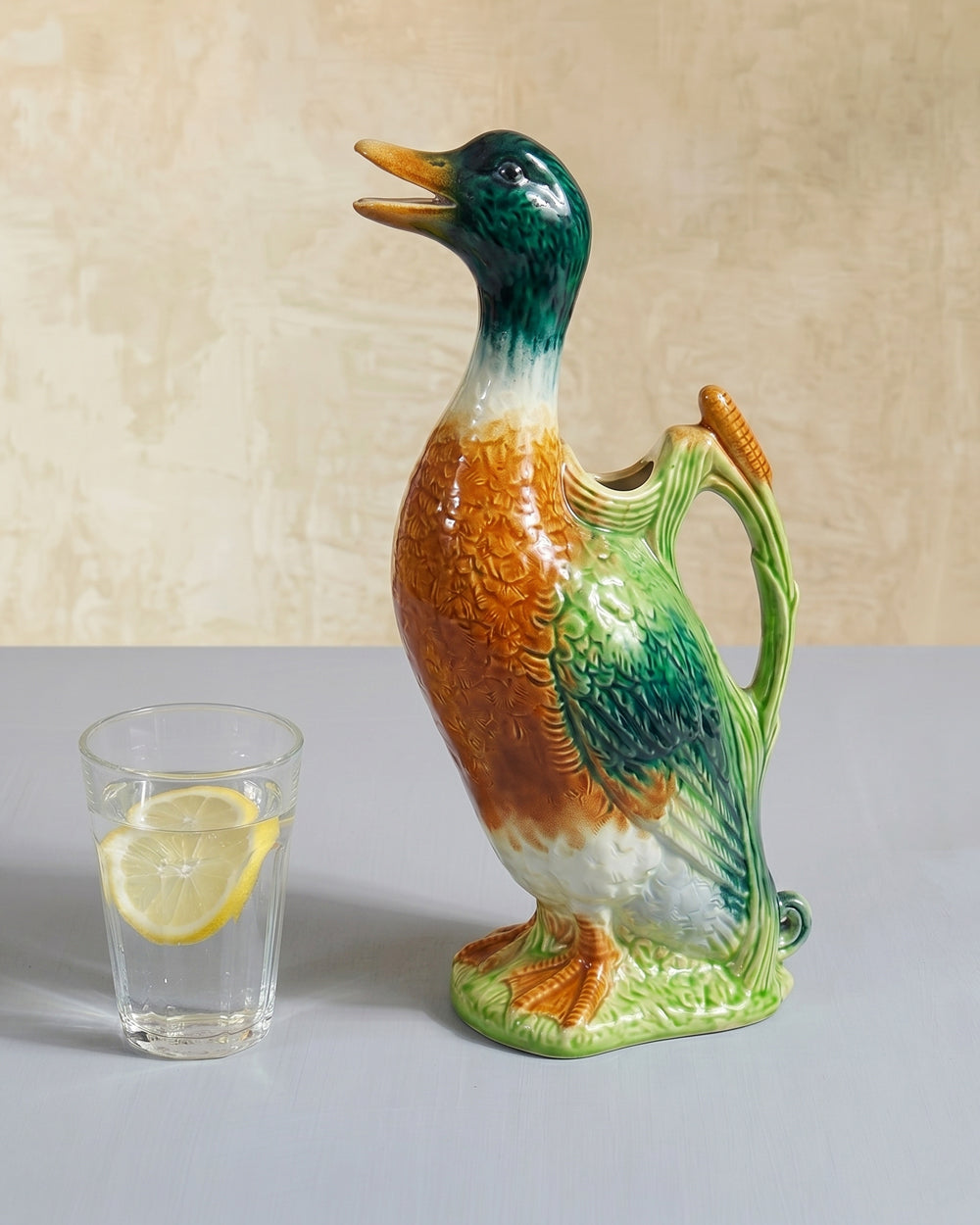 Vintage French ceramic duck water pitcher figurine by St. Clément with colorful glazed detailing next to a glass of water with a lemon slice, displayed on a plain background, available at Wildschut Antiques.