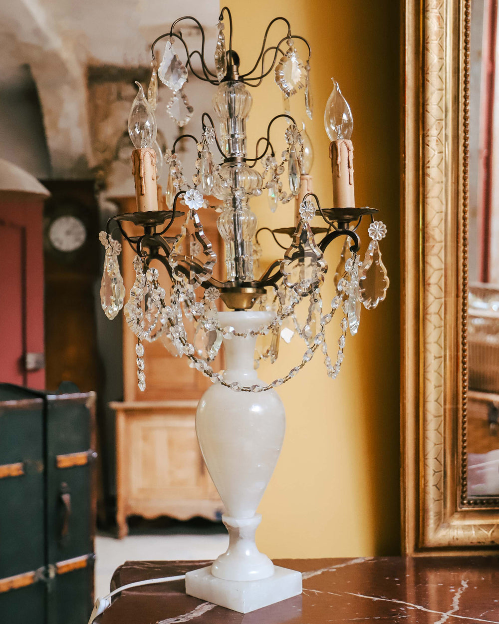 A 19th C French crystal chandelier-style table lamp with a white base, featuring a central candle holder and decorative crystal elements, displayed in front of an antique golden mirror with antique armoires in the background.