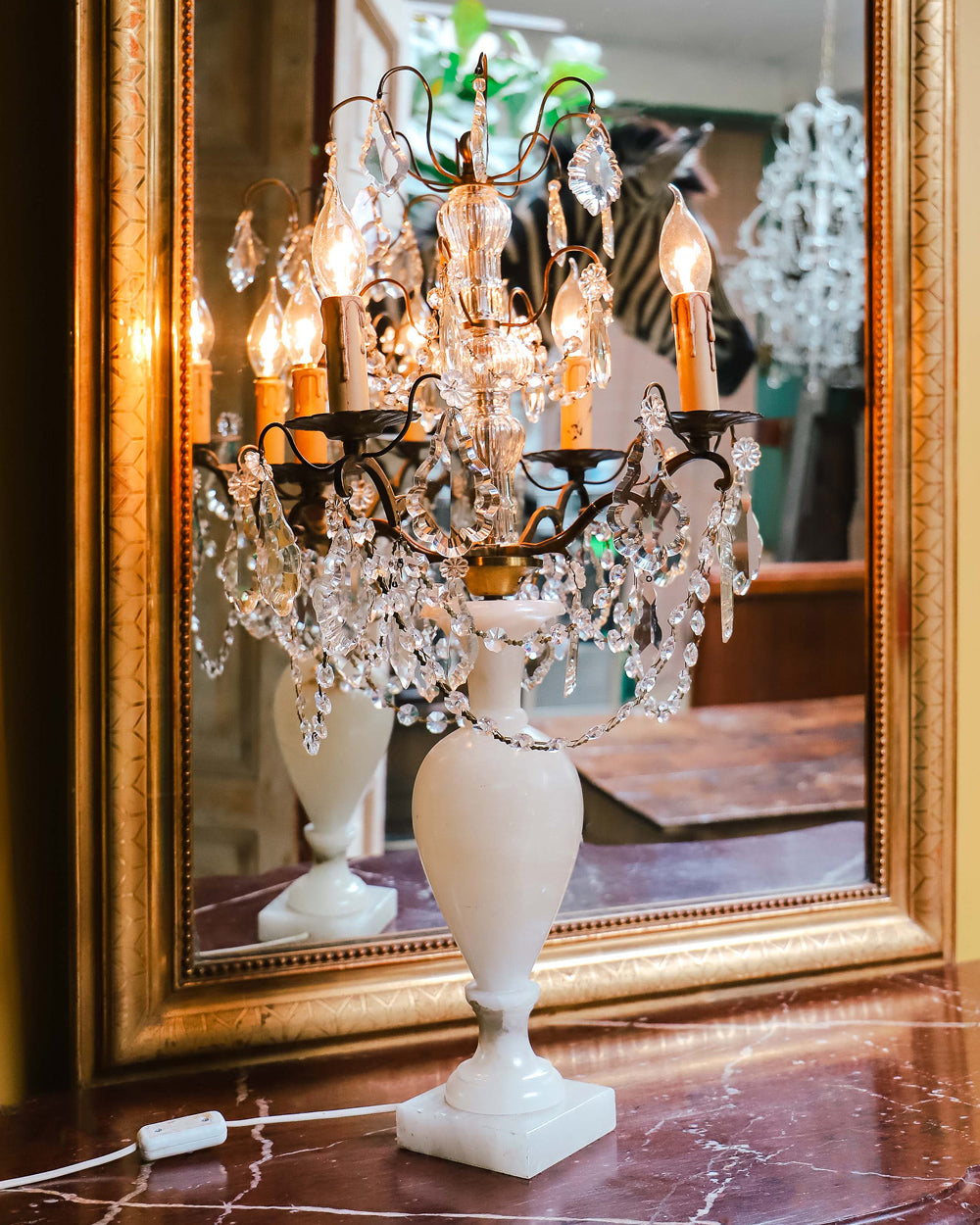 A 19th C French crystal chandelier-style table lamp with a white base, featuring a central candle holder and decorative crystal elements, displayed in front of an antique golden mirror.