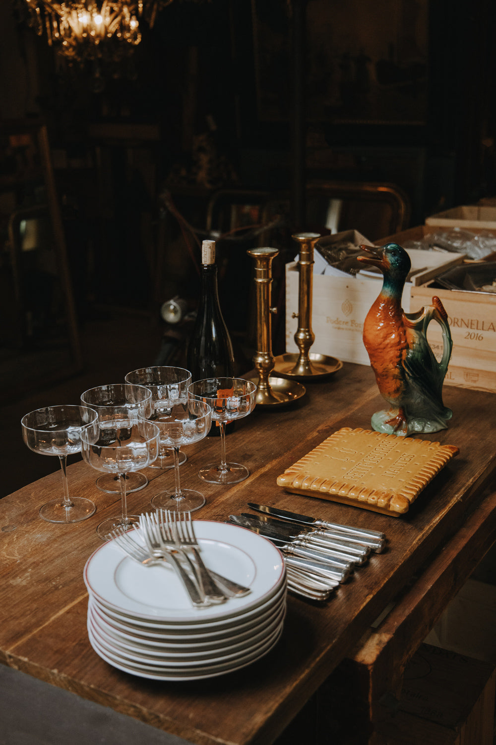 Table setting in the Wildschut Antiques warehouse featuring Art Deco-style champagne coupes, two large brass candle holders, a St. Clement ceramic duck pitcher, a set of porcelain plates, silver-plated cutlery, and a bottle of wine—all bathed in a warm, moody light.