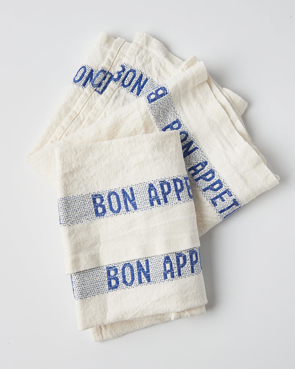 Folded white linen dish towels with 'BON APPÉTIT' embroidered in blue along the edges, by Charvet Editions, available at Wildschut Antiques