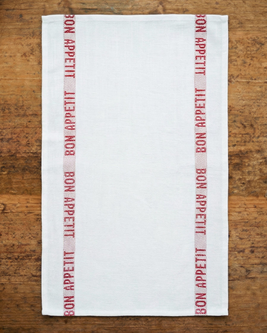 White linen towel with red 'Bon Appetit' text on a wooden surface, by Charvet Editions available via Wildschut Antiques.