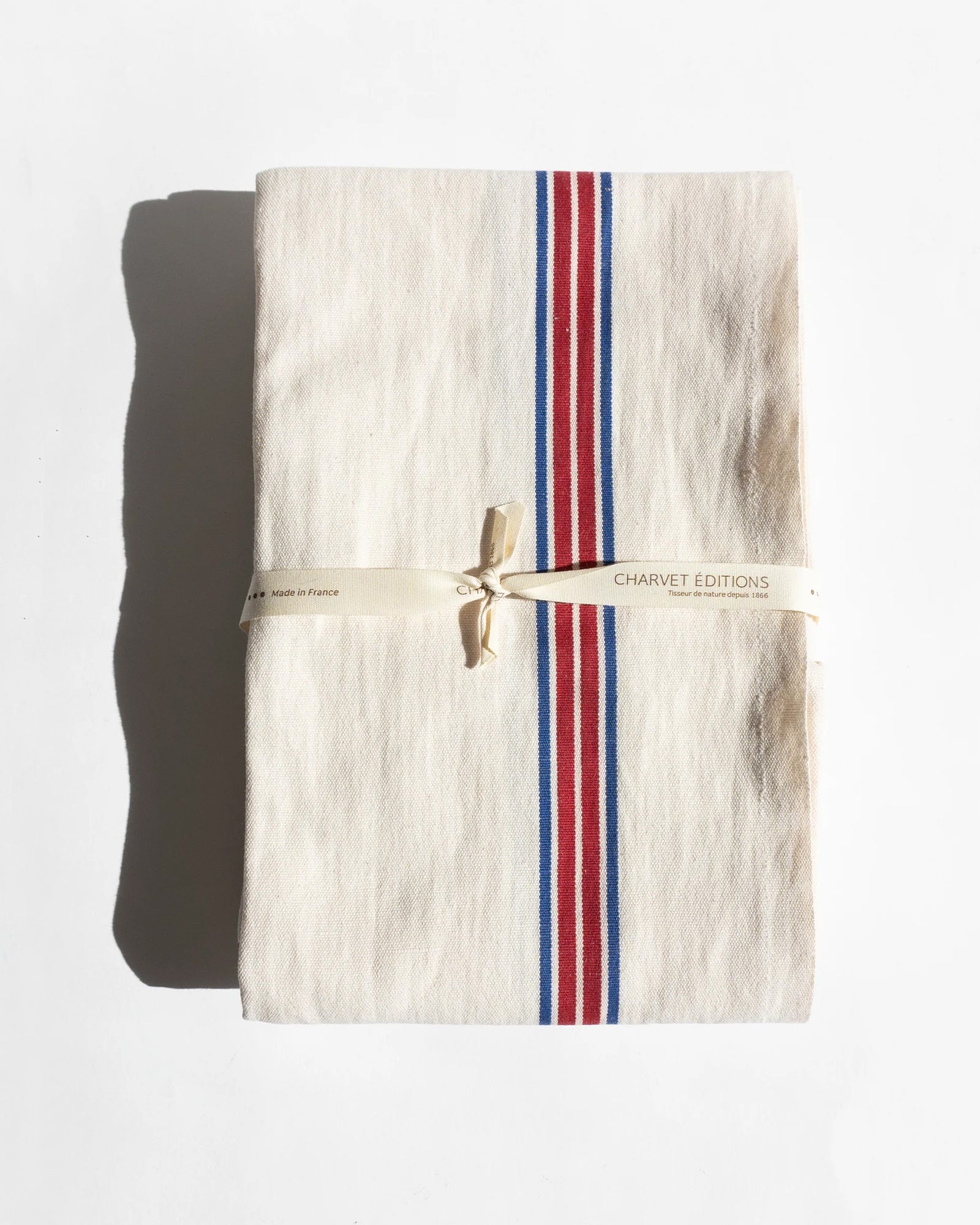 A folded white dish towel with blue and red stripes on the sides by Charvet Editions available at Wildschut Antiques