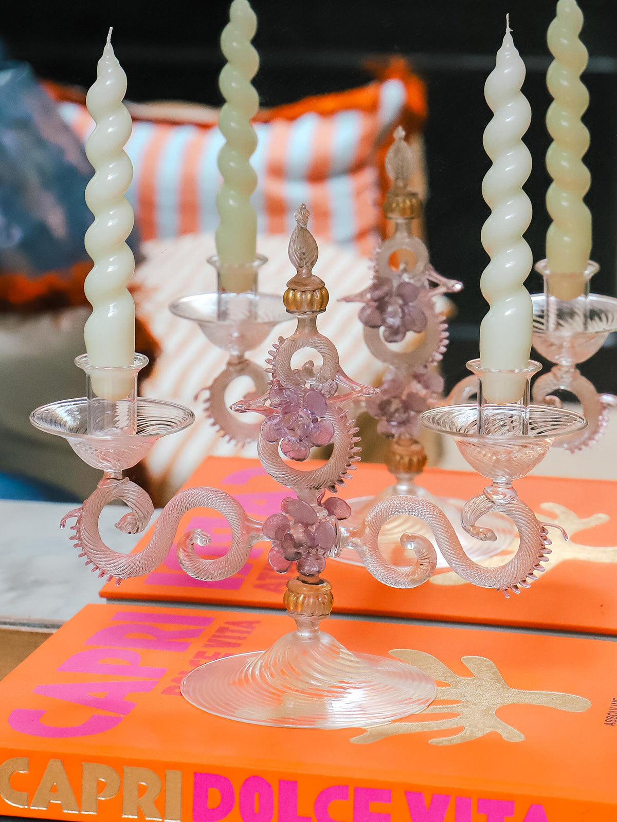 Venetian Murano glass candleholder holding two cream-white swirl candles, set atop the Assouline book *Capri Dolce Vita* and positioned before an antique gilded mirror reflecting the candleholder and a cluster of colorful striped pillows—all at Wildschut Antiques.