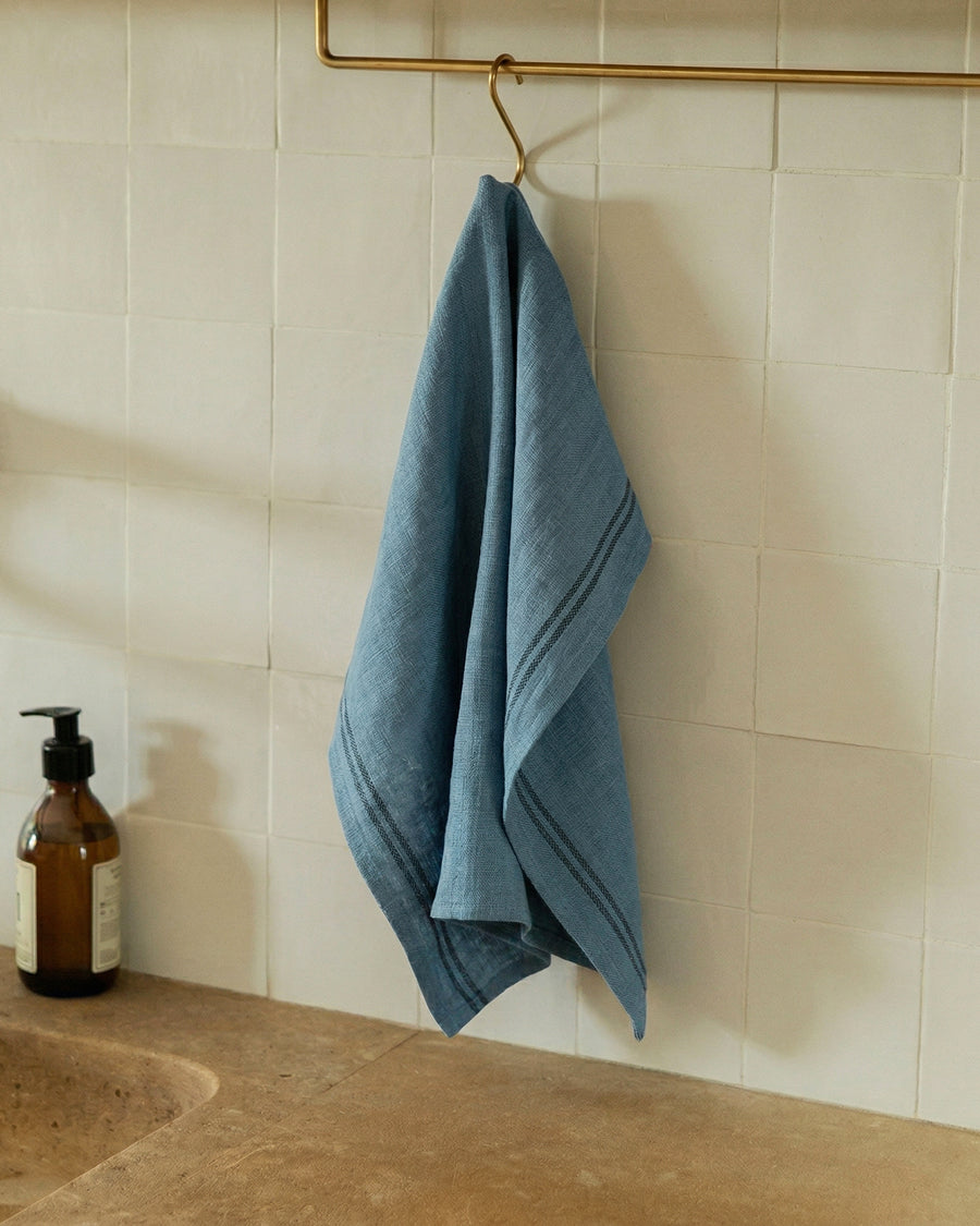 Blue linen towel hanging on a gold hook against a tiled wall next to a a bottle of soap. Linen country towel by Charvet Editions available via Wildschut Antiques.