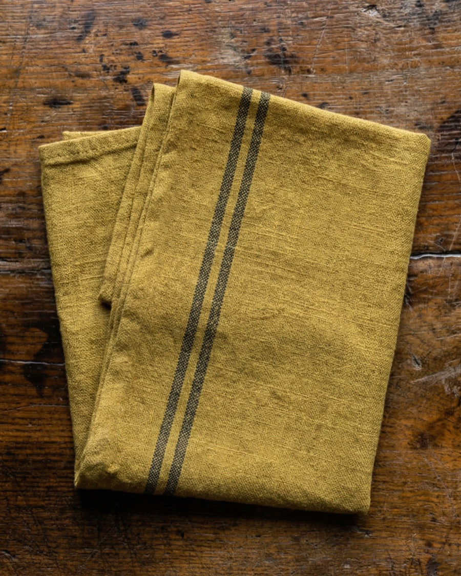 Folded ochre yellow linen towel with dark stripes on a wooden surface. Linen country towel by Charvet Editions available via Wildschut Antiques.