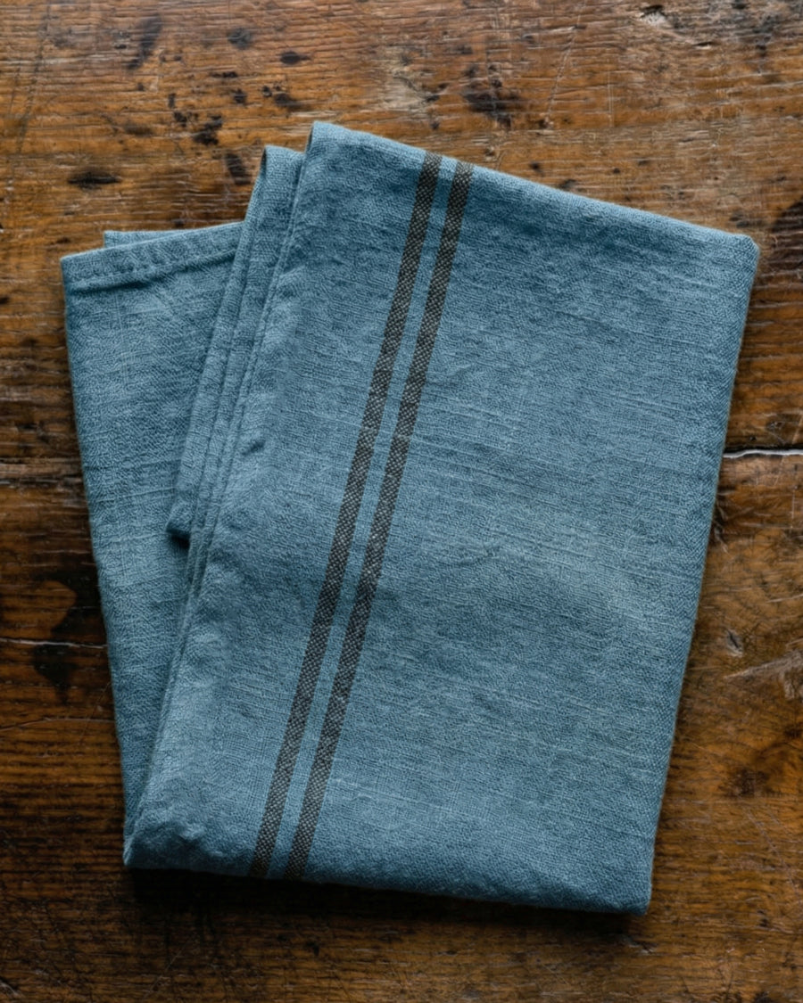 Folded linen blue towel with dark stripes on a wooden surface. Linen country towel by Charvet Editions available via Wildschut Antiques.
