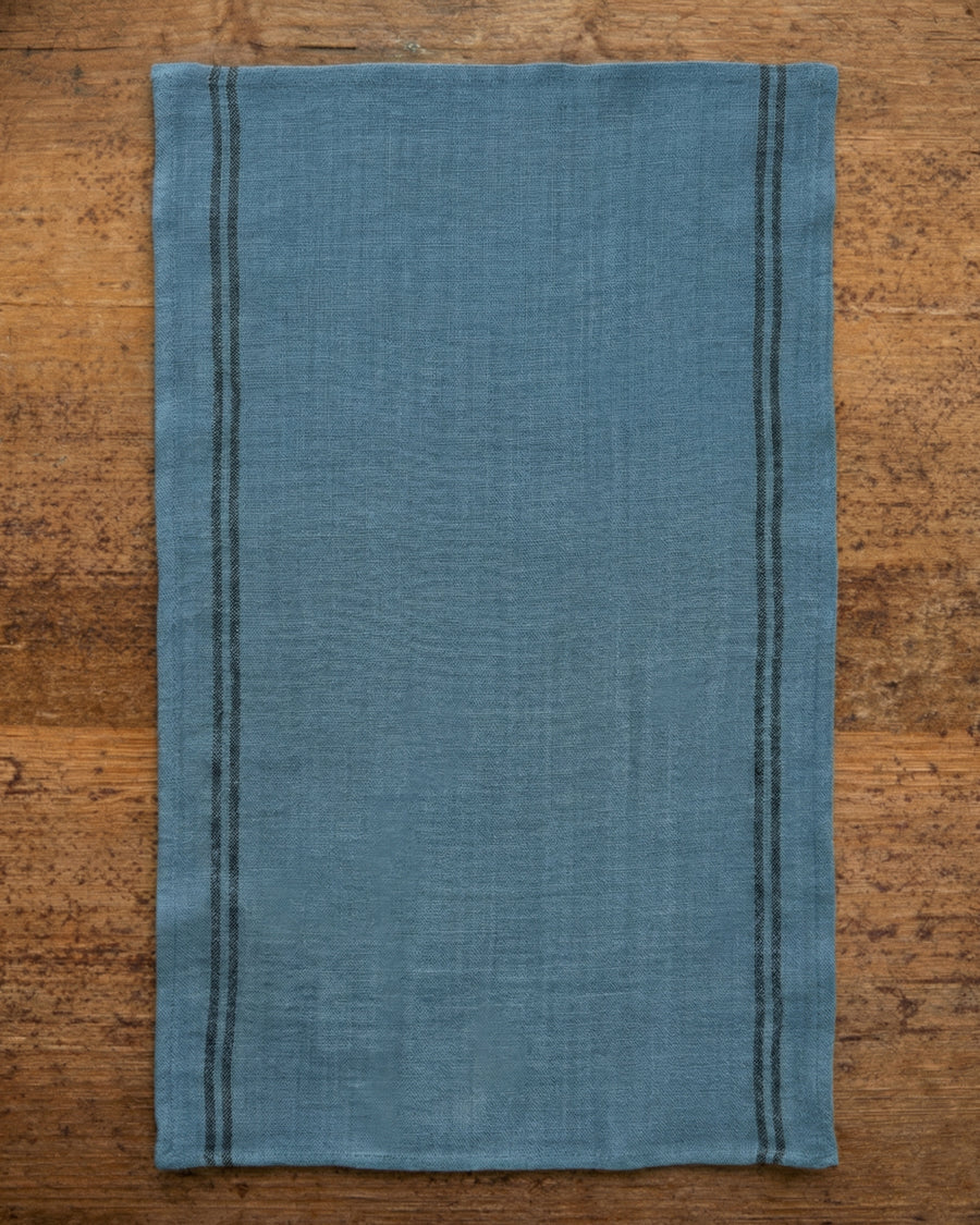 Blue linen towel with dark stripes on a wooden surfaceLinen country towel by Charvet Editions available via Wildschut Antiques.