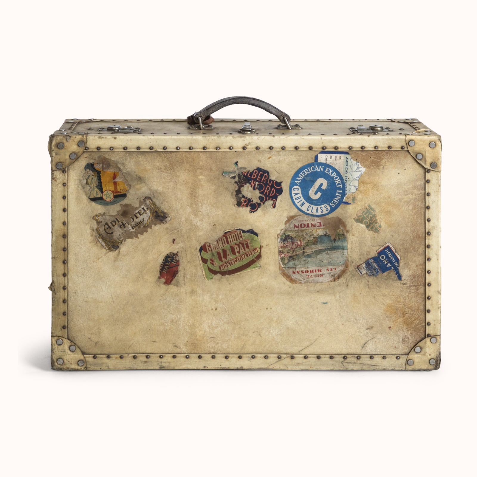 A vintage French cream vellum white leather suitcase or valise with brass studs and lock plates, adorned with various travel labels and stickers, featuring a handle and a lock, available at Wildschut Antiques