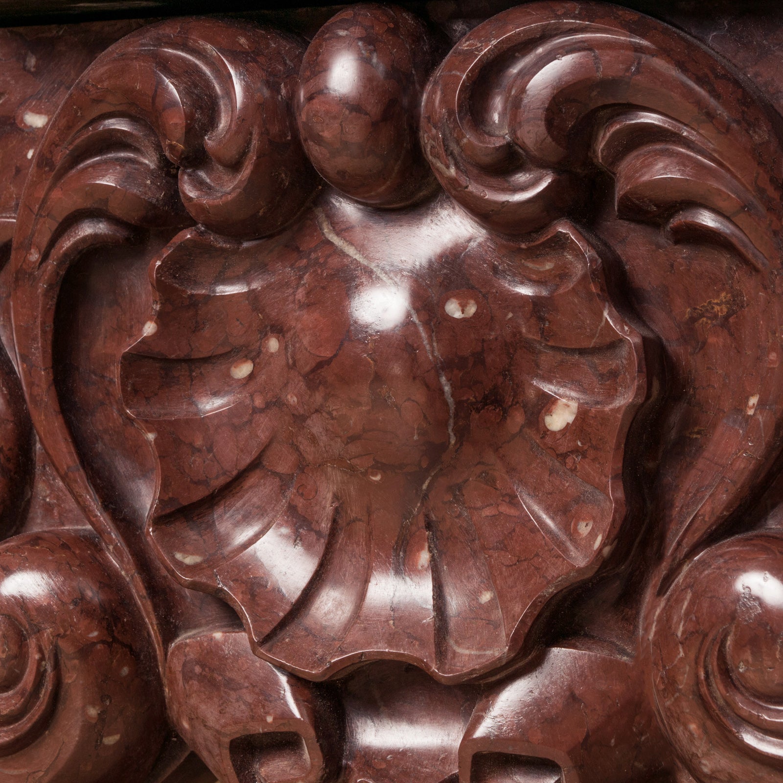 Close-up of a shell shape at the front of a grand antique French Rococo Louis XV fireplace mantel, crafted from richly veined red-brown marble.