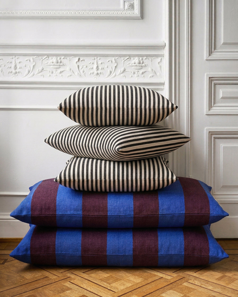 Stack of striped pillows on a wooden floor with decorative wall in the background, available via Wildschut Antiques.