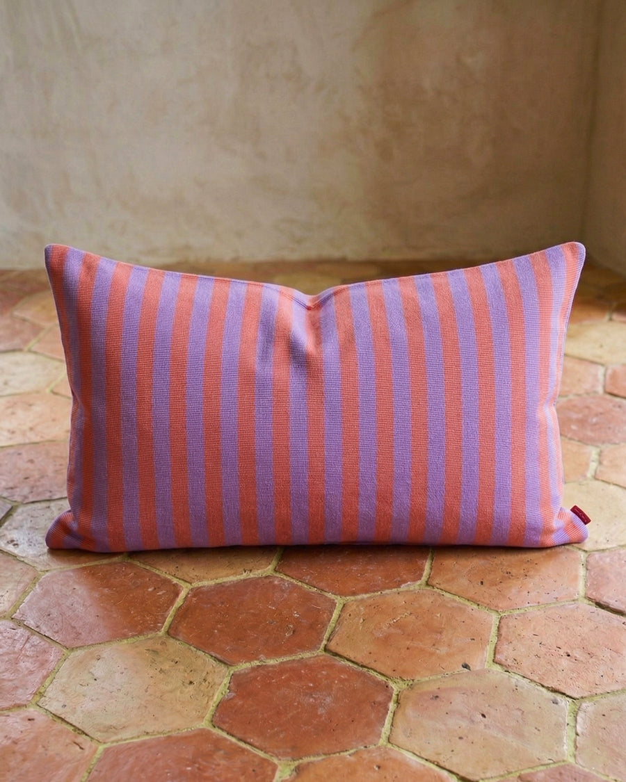 Lila and pink striped pillow on a terracotta-tiled floor with a textured wall background, available via Wildschut Antiques.