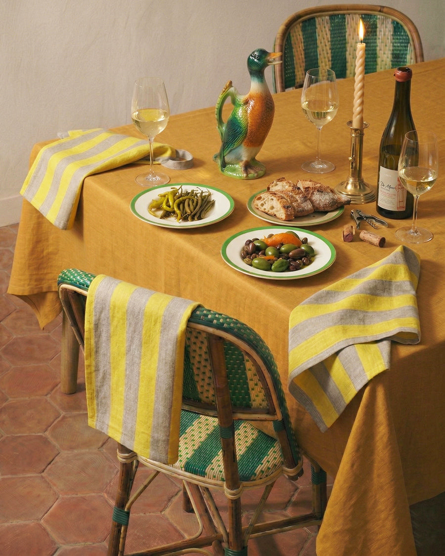 Dining table set with food, wine, and decorative items such as an antique French brass candle holder and St. Clement French ceramic duck Pitcher, on a yellow tablecloth. Available via Wildschut Antiques.