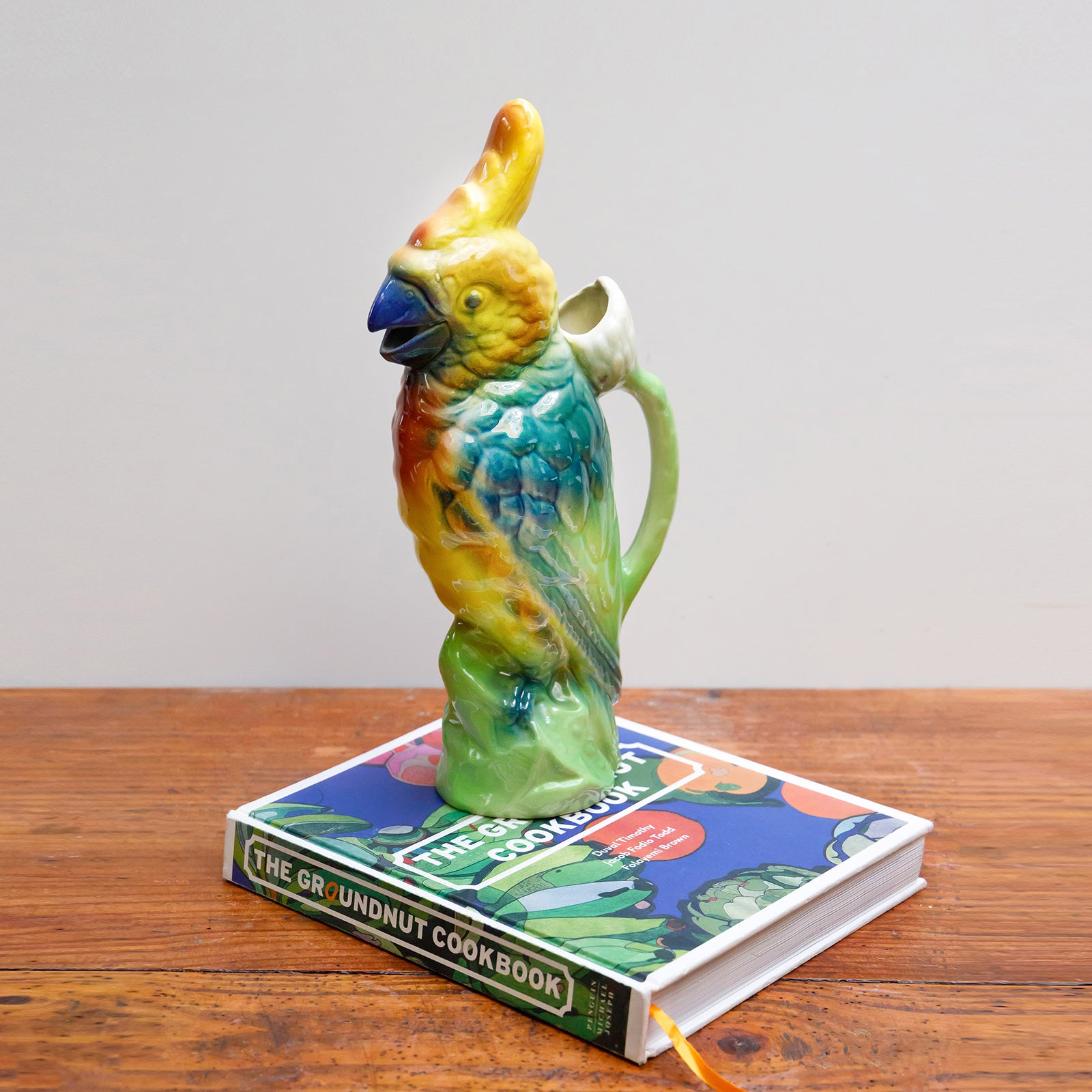 A vibrantly colored ceramic pitcher in the shape of a cockatoo with a green body and yellow details by St. Clement, France, displayed on a colourful cookbook on a wooden table