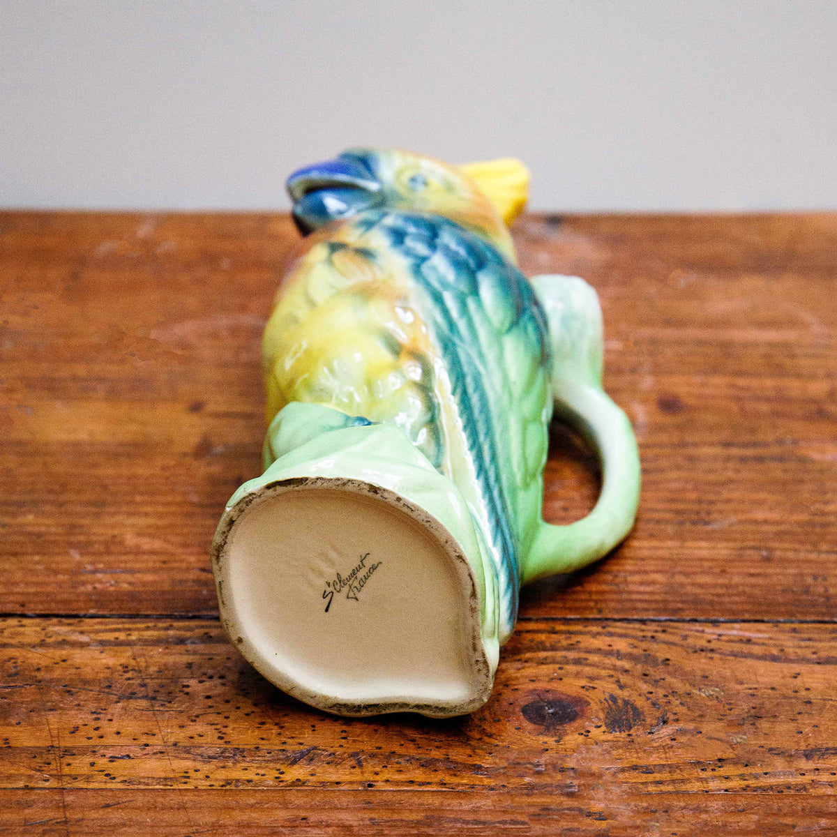 Bottom view of a vibrantly colored ceramic pitcher in the shape of a cockatoo with a green body and yellow details by St. Clement, France, displayed lying on a wooden table