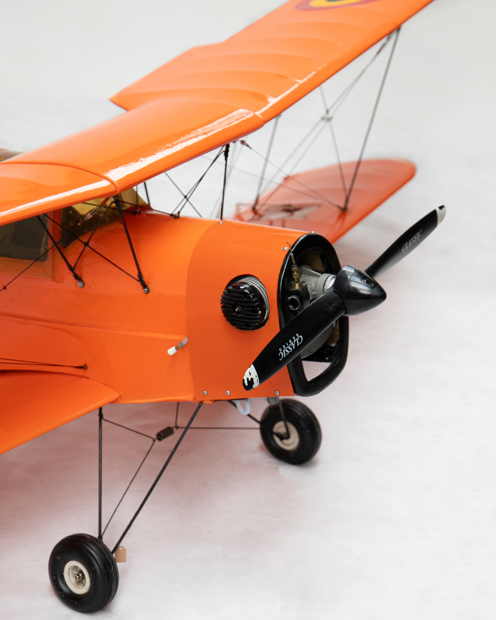 Close-up of an orange biplane model with a black propeller and black wheels, photographed on a light gray studio background.