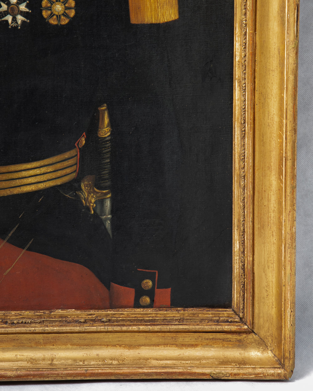 Close-up detail of a framed painting showing part of a military uniform with gold epaulets, a sword hilt, and a red coat against a dark background, with the ornate gilded frame prominently visible.