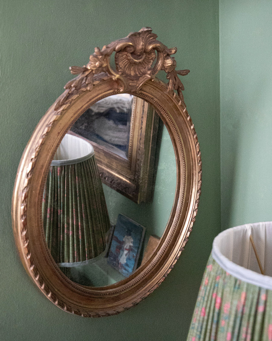 An ornate antique French oval wall mirror with a carved gold frame hangs on a sage green wall, reflecting a pleated green lampshade and a framed picture.