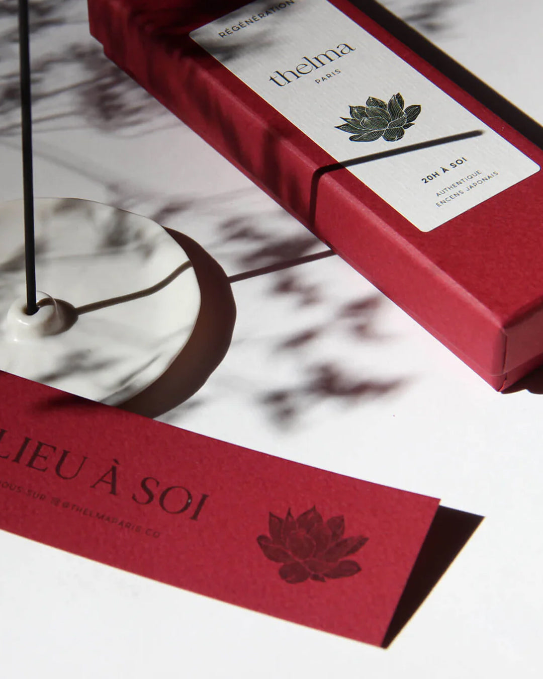 Two red boxes of Thelma Paris incense sticks with white labels featuring a cherry blossom. A white incense holder and a smokeless incense stick are also visible, placed on a surface with a shadow falling over them.