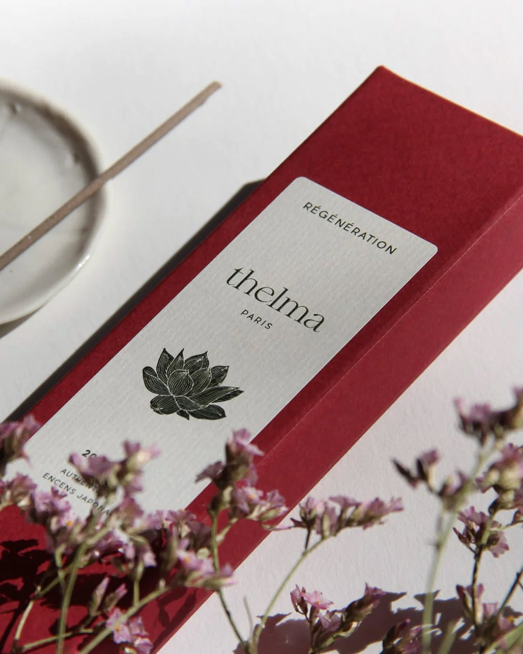 A red box of Thelma Paris incense sticks with a white label featuring a cherry blossom. A white incense holder and a smokeless incense stick are also visible, placed on a surface with a small purple flowers.