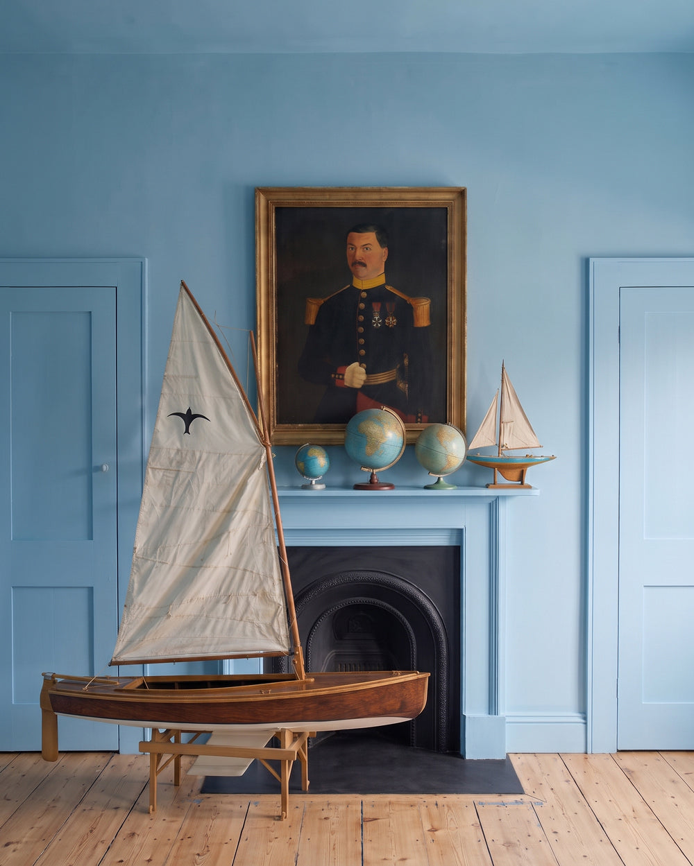 A styled interior scene with a large wooden Valk model sailboat in the foreground, a blue fireplace mantel with globes and a framed portrait, and blue walls and doors in the background.