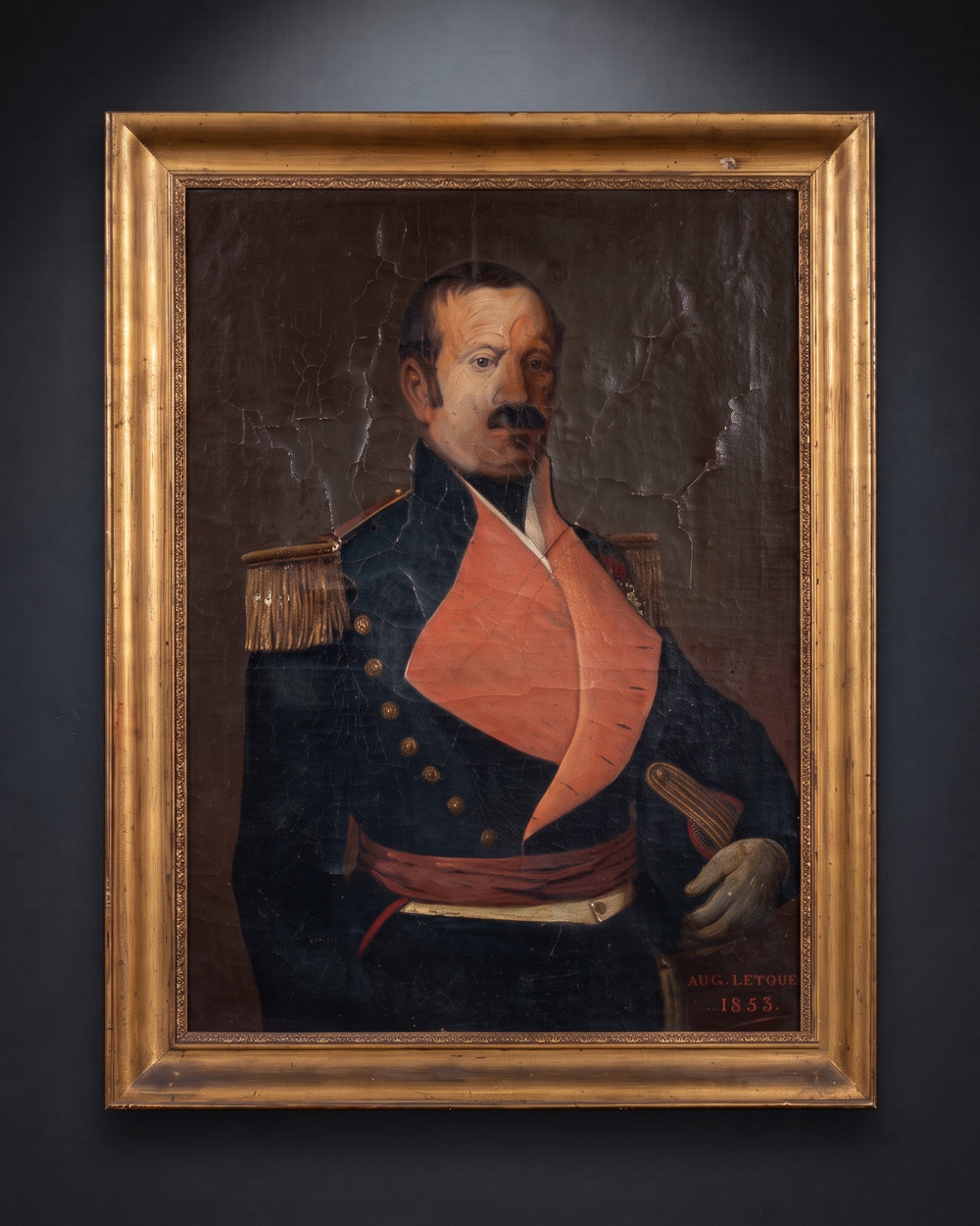 A framed historical antique portrait oil painting of a man in a dark military uniform with a bright orange-red coat and gold epaulettes, set in a wide ornate gold frame against a dark wall. Available via Wildschut Antiques.