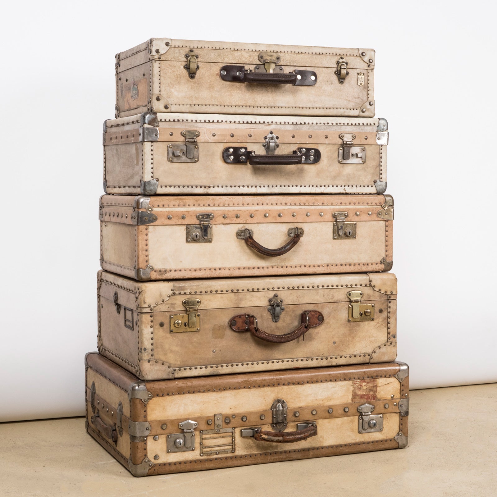 Elegant set of five early 20th-century white leather, vellum, or parchment suitcases, each featuring original travel labels, displayed against a white wall. Available at Wildschut Antiques.