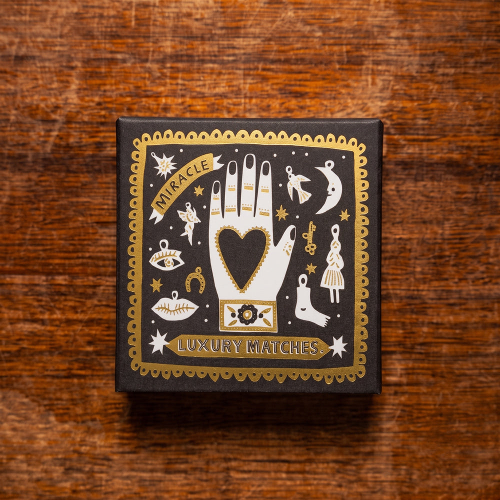 A square black matchbox of 'Miracle Luxury Matches' with ornate gold foil illustrations of a hand, stars, and mystical symbols, placed on a rustic wooden surface. Design by Archivist Gallery. Available at Wildschut Antiques & Decor.