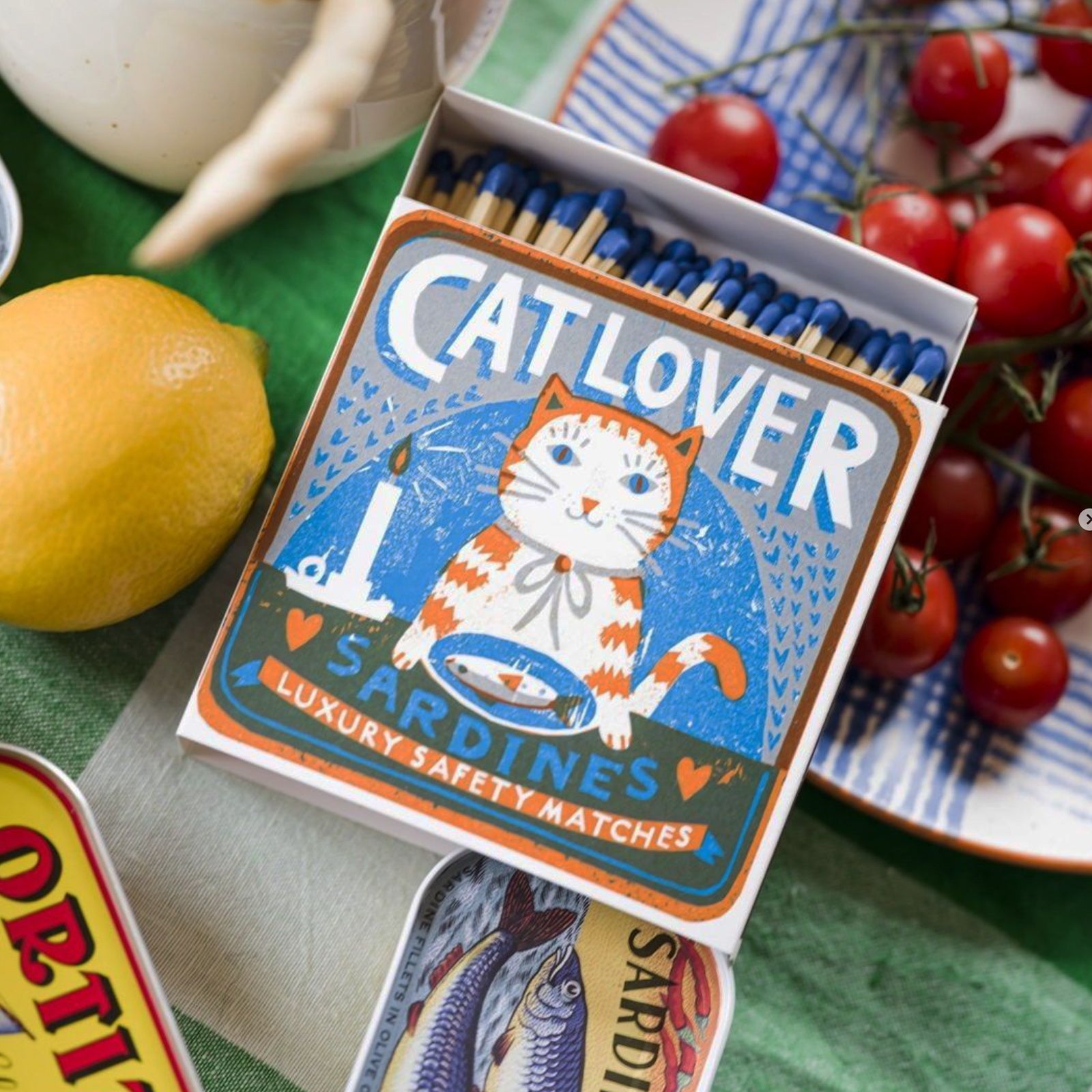 Matches box labeled 'Cat Lover' with a cat illustration on a table with a lemon, a can of sardines, and tomatoes.