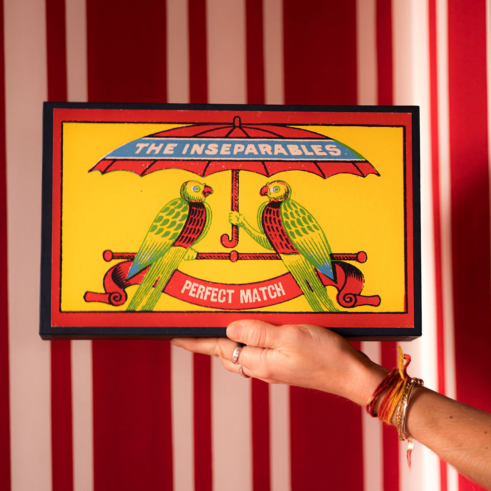 Giant Matchbox decorated with two birds under an umbrella and text 'The Inseparables Perfect Match' against a red and white striped background. Design by Archivist Gallery, available at Wildschut Antiques.