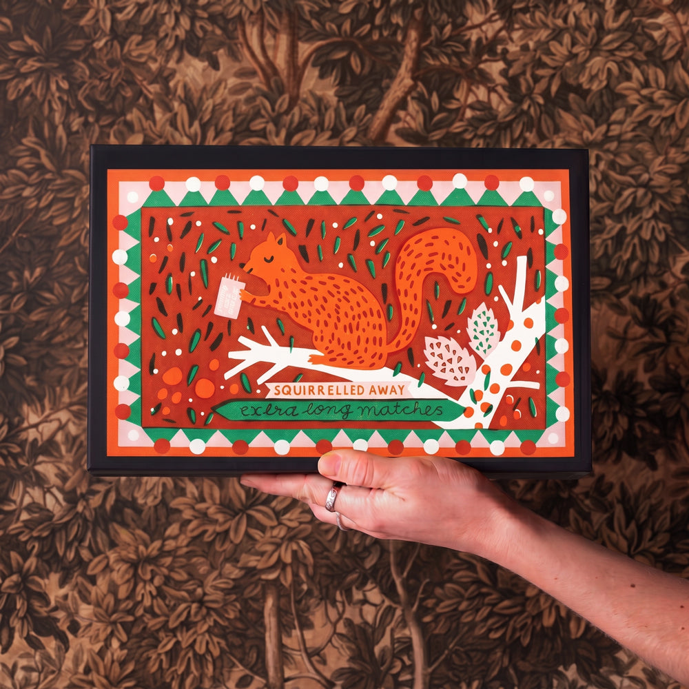 A hand holds a large matchbox featuring a print of a stylized orange squirrel on a branch with the text 'SQUIRRELLED AWAY' and 'extra long matches'. The print has a black frame and is set against a brown patterned background.