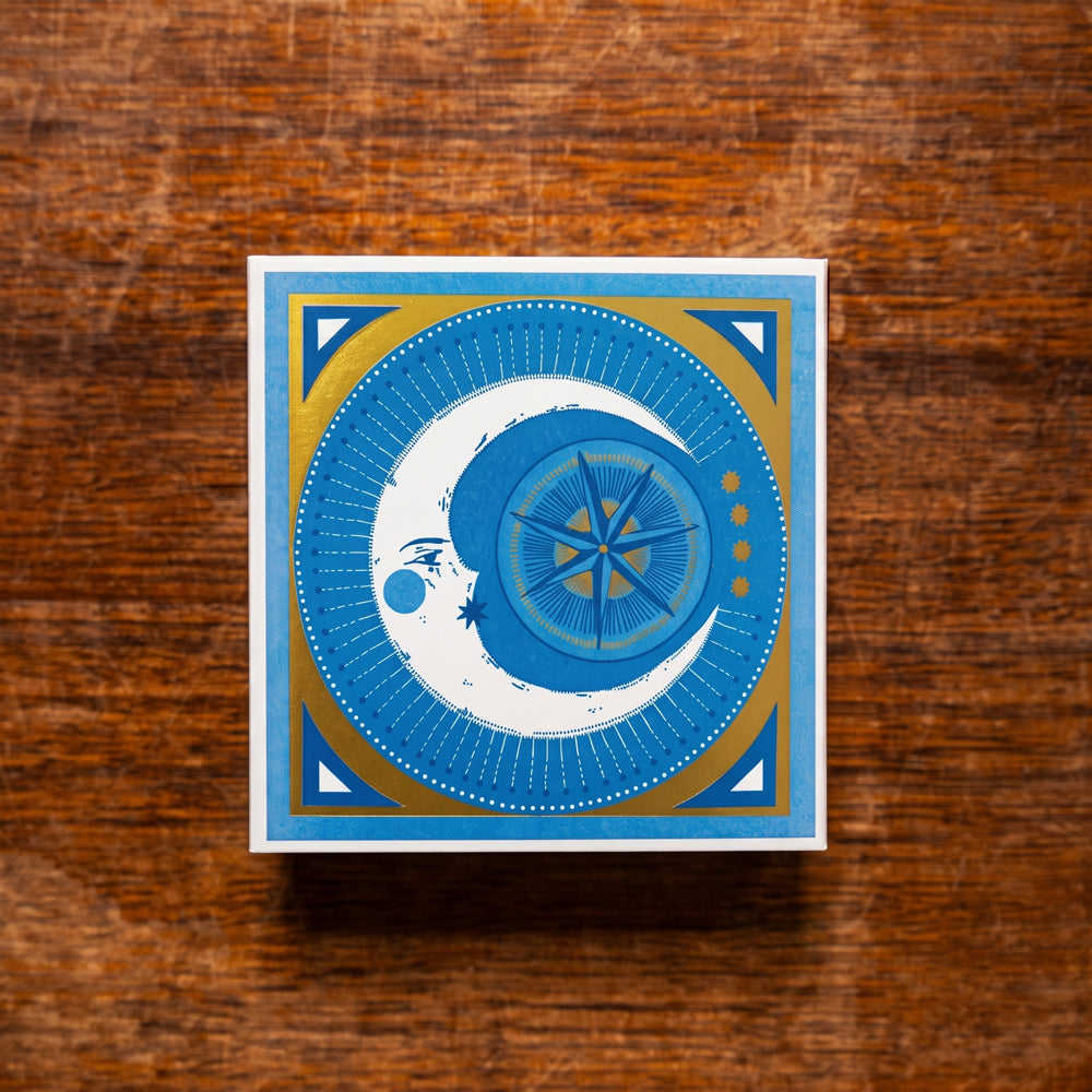 A square decorative matchbox with an illustrated celestial design in blue, white, and gold, placed on a rustic brown wooden surface.