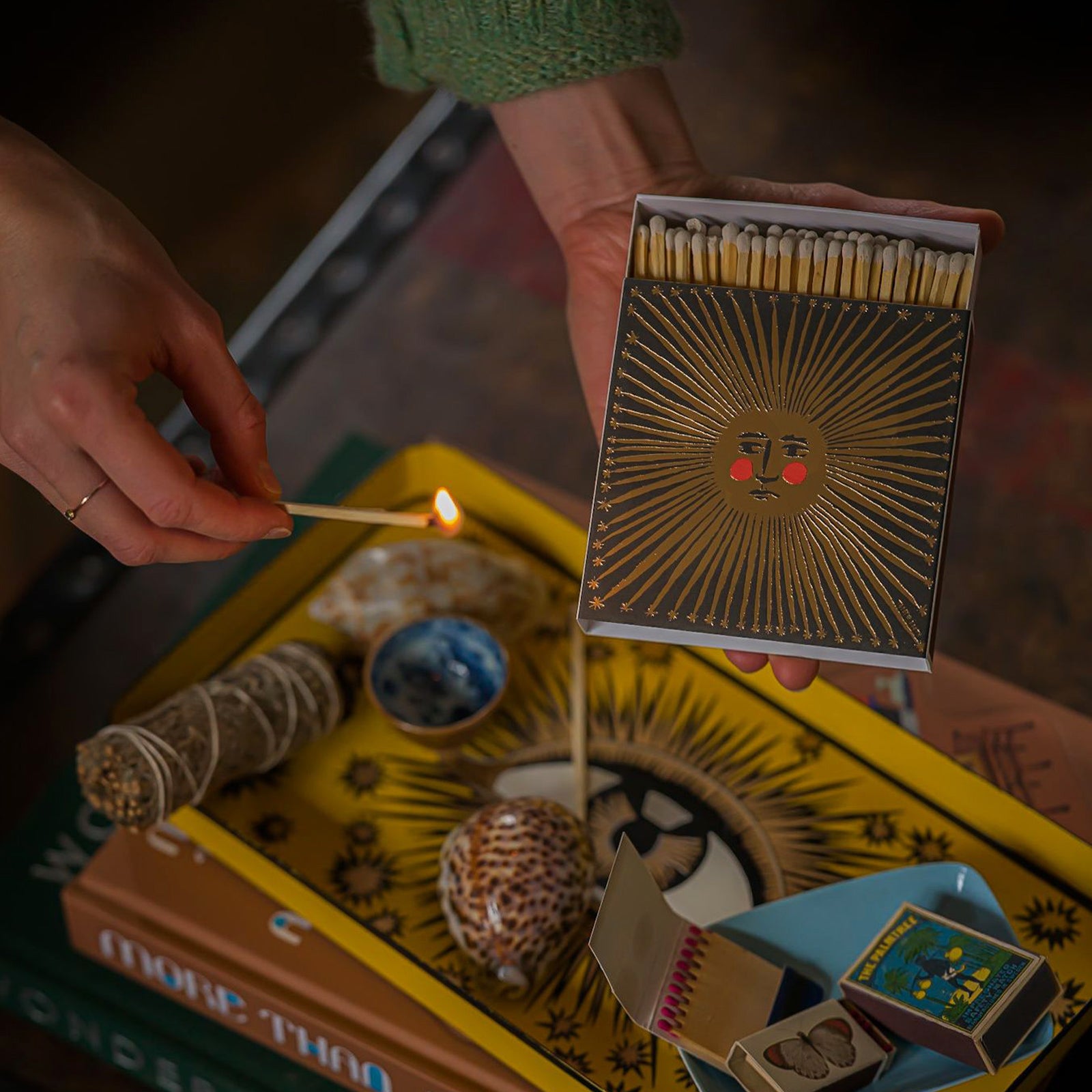 Person lighting a match from a decorative matchbox with a golden sun design on a table with books and other items.